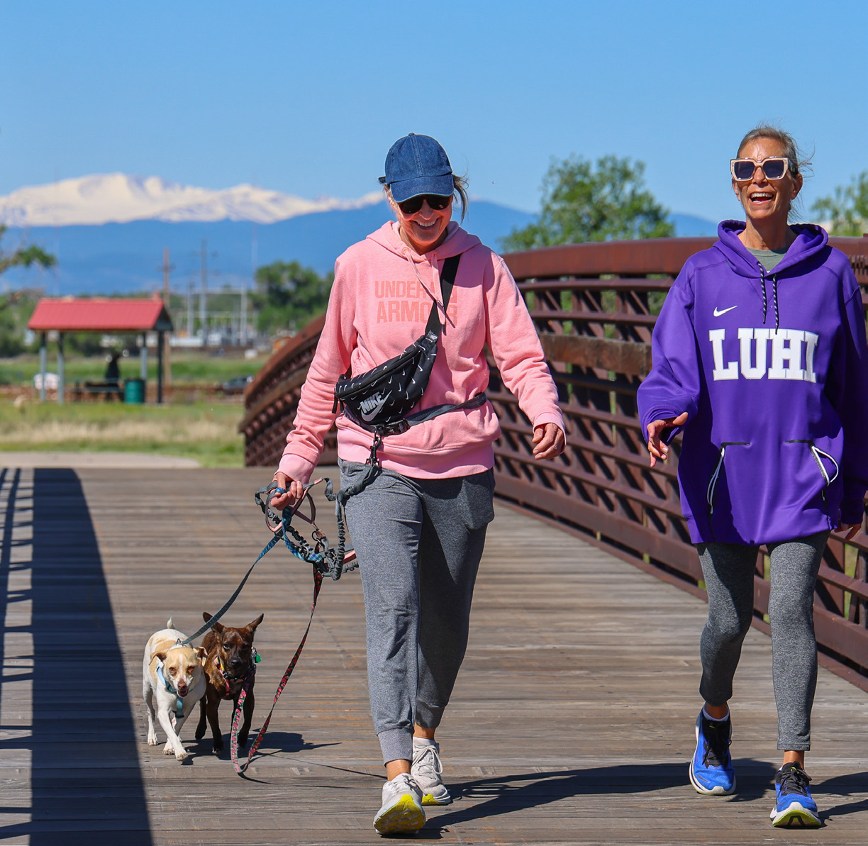 Two women walking their dogs over a bridge on a nice day. Mountain and blue sky in background.