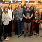 A group of twelve people in business attire smiling, one holding an award plaque.