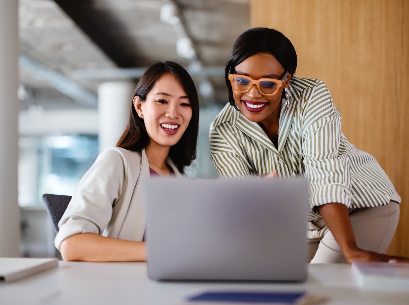 Two professional women discussing work on a laptop, demonstrating teamwork and collaboration in a modern office setting.