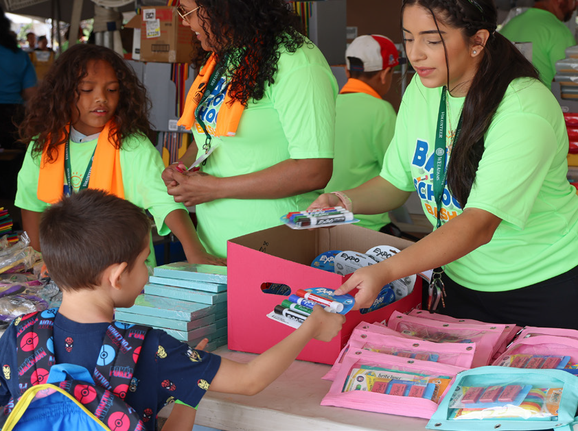 Children receiving school supplies at Back-to-School event, guided by volunteers in bright shirts.