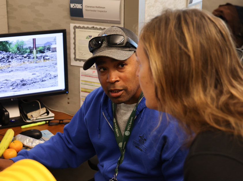 Clarence Hollimon, stormwater inspector, engaged in conversation, desk with paperwork, monitor displaying construction site image.