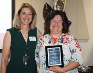 Two women smiling; one holds a plaque recognizing Amy Clement for outstanding contributions.