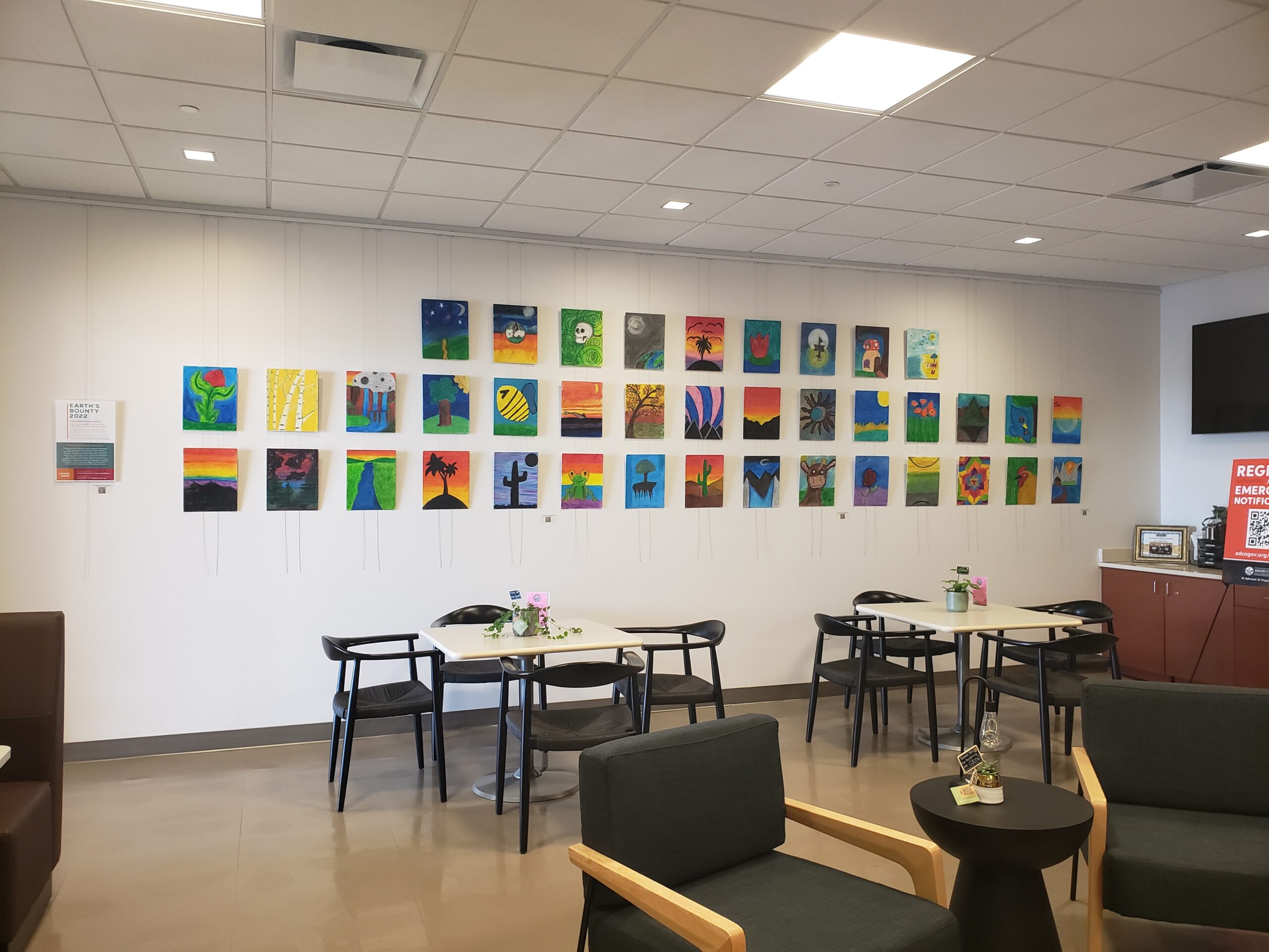 Art exhibit with colorful paintings on a white wall, tables and chairs below.