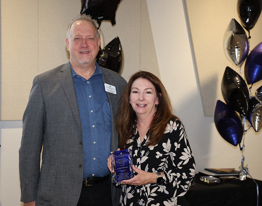 Man and woman smiling; woman holding an award, black and silver balloons in background