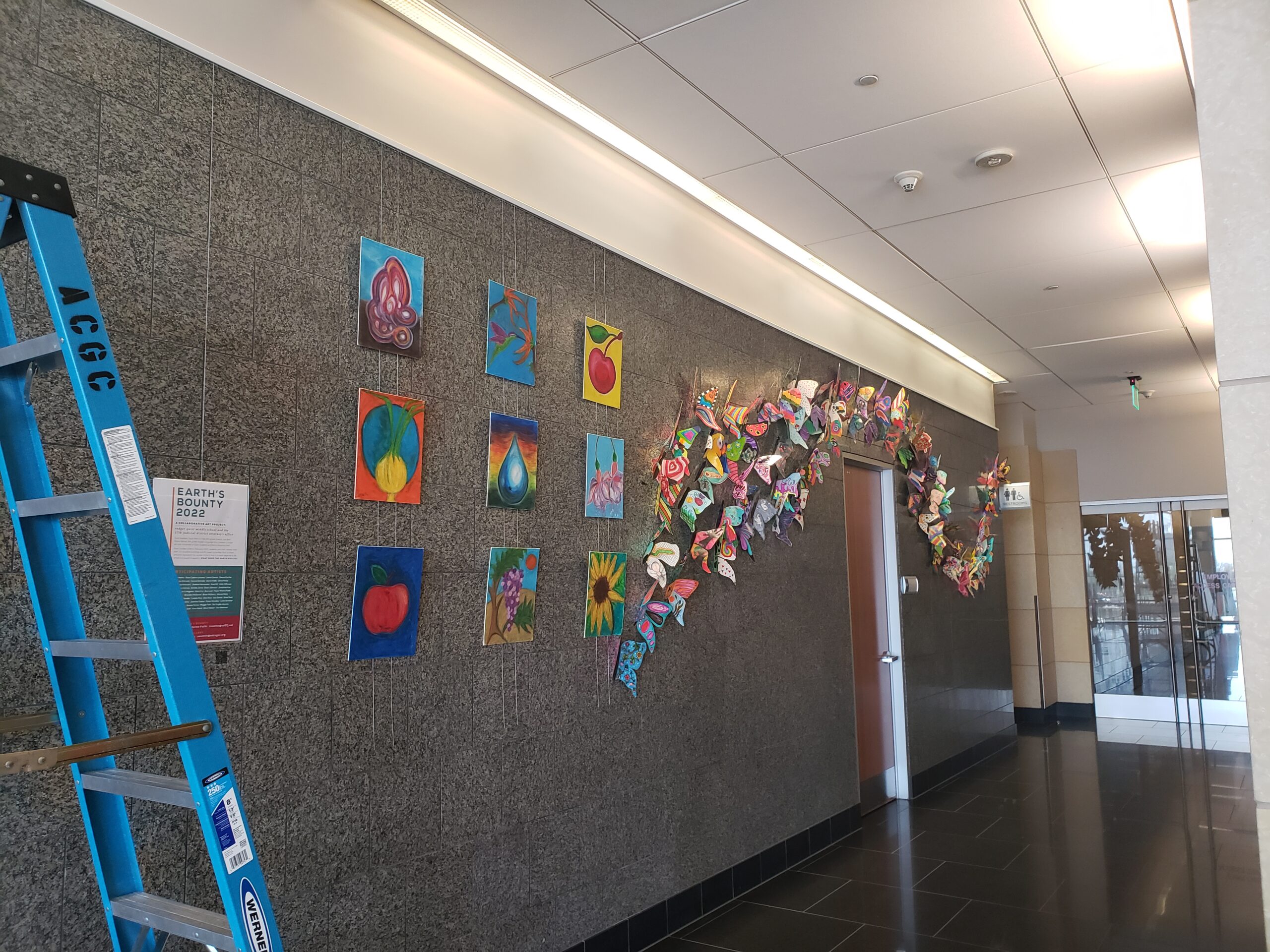 Colorful art display with vibrant paintings and butterfly sculpture, titled "Earth's Bounty 2022," inside a modern building.