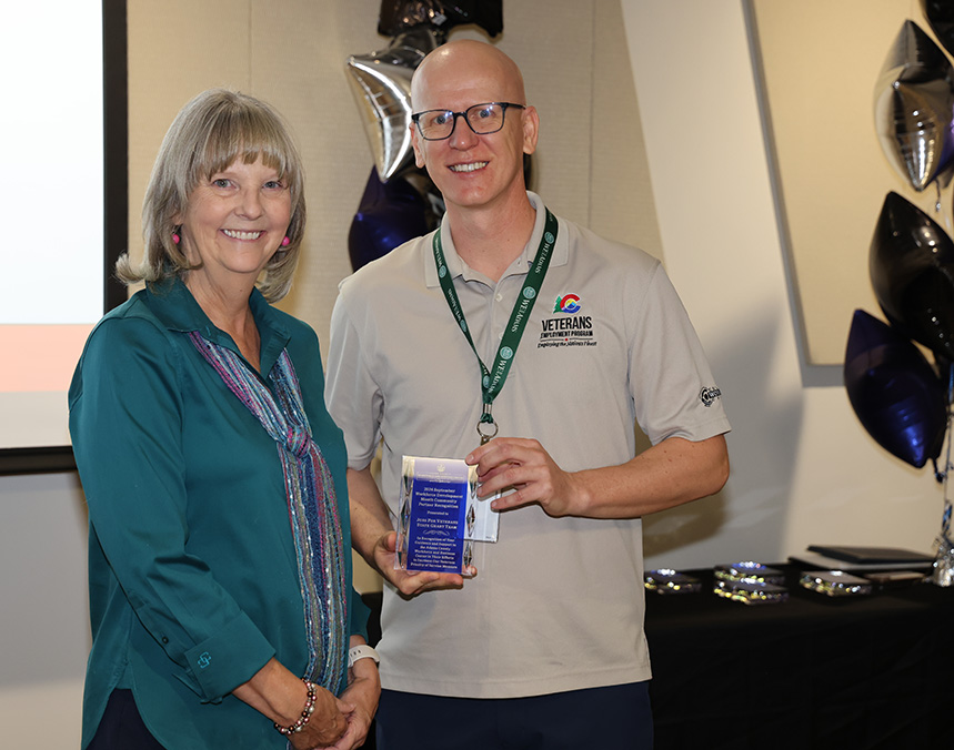 Woman and man smiling, holding award; man wears "Veterans Upward Bound" shirt.
