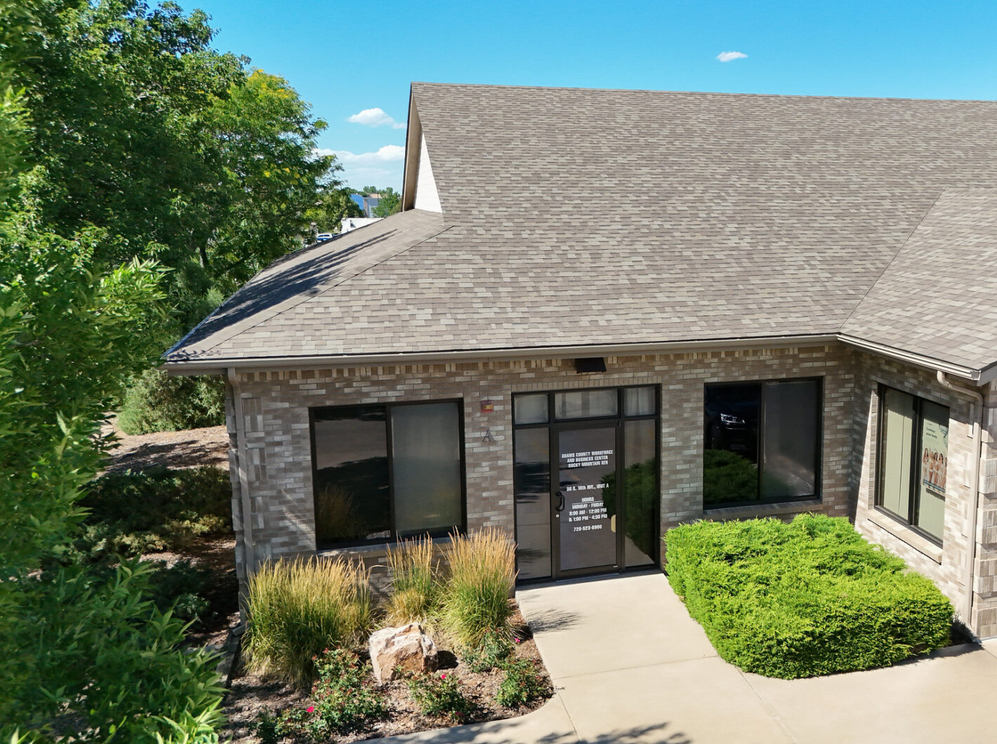 Grey brick office building with landscaped entrance, green foliage, and clear glass doors displaying hours and contact information.