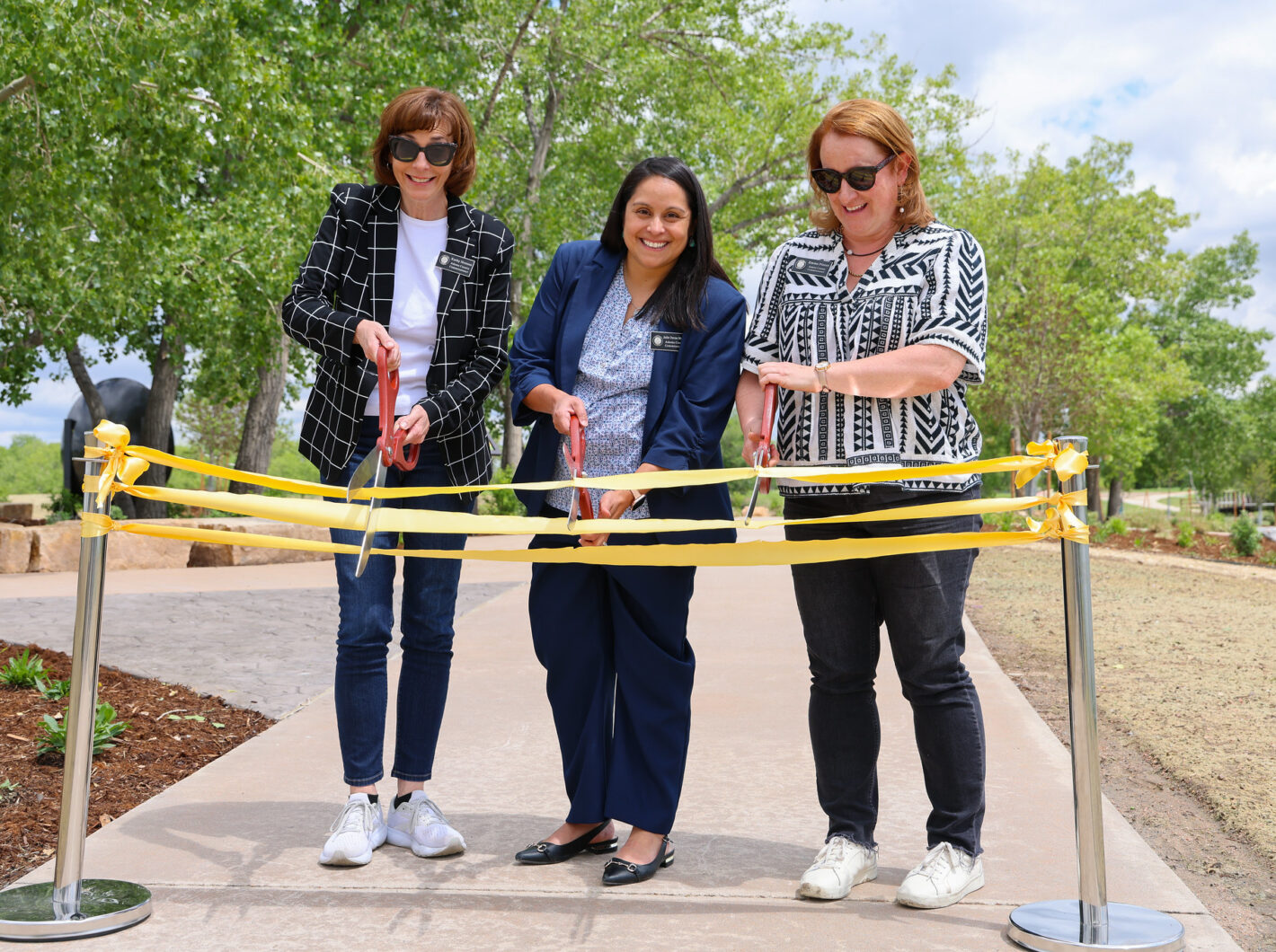Three women, including the central figure, smiling while cutting a yellow ribbon at an outdoor ceremony.