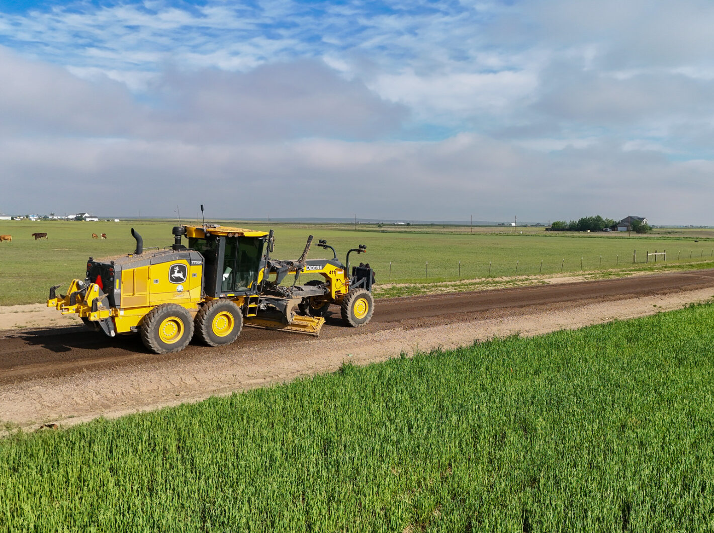 Yellow road grader on a rural dirt road, green fields surrounding.
