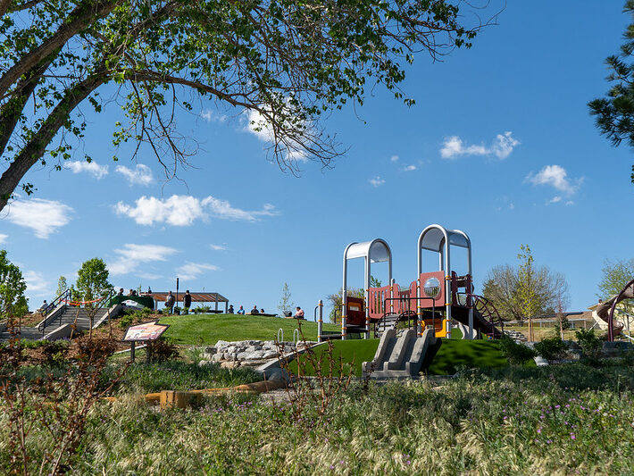 Playground surrounded by green grass, trees, and people under a blue sky