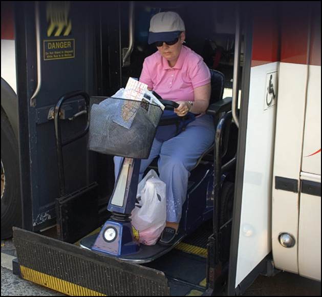 A person on a mobility scooter boards a bus using a lift; wearing a pink shirt and cap.