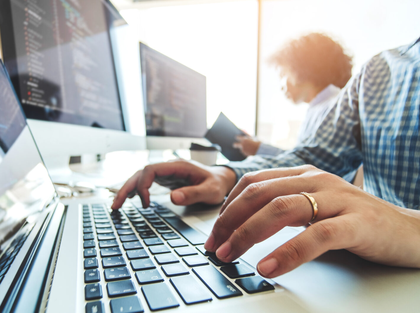 Hands typing on a laptop keyboard, with programming code displayed on multiple monitors in a bright office environment.