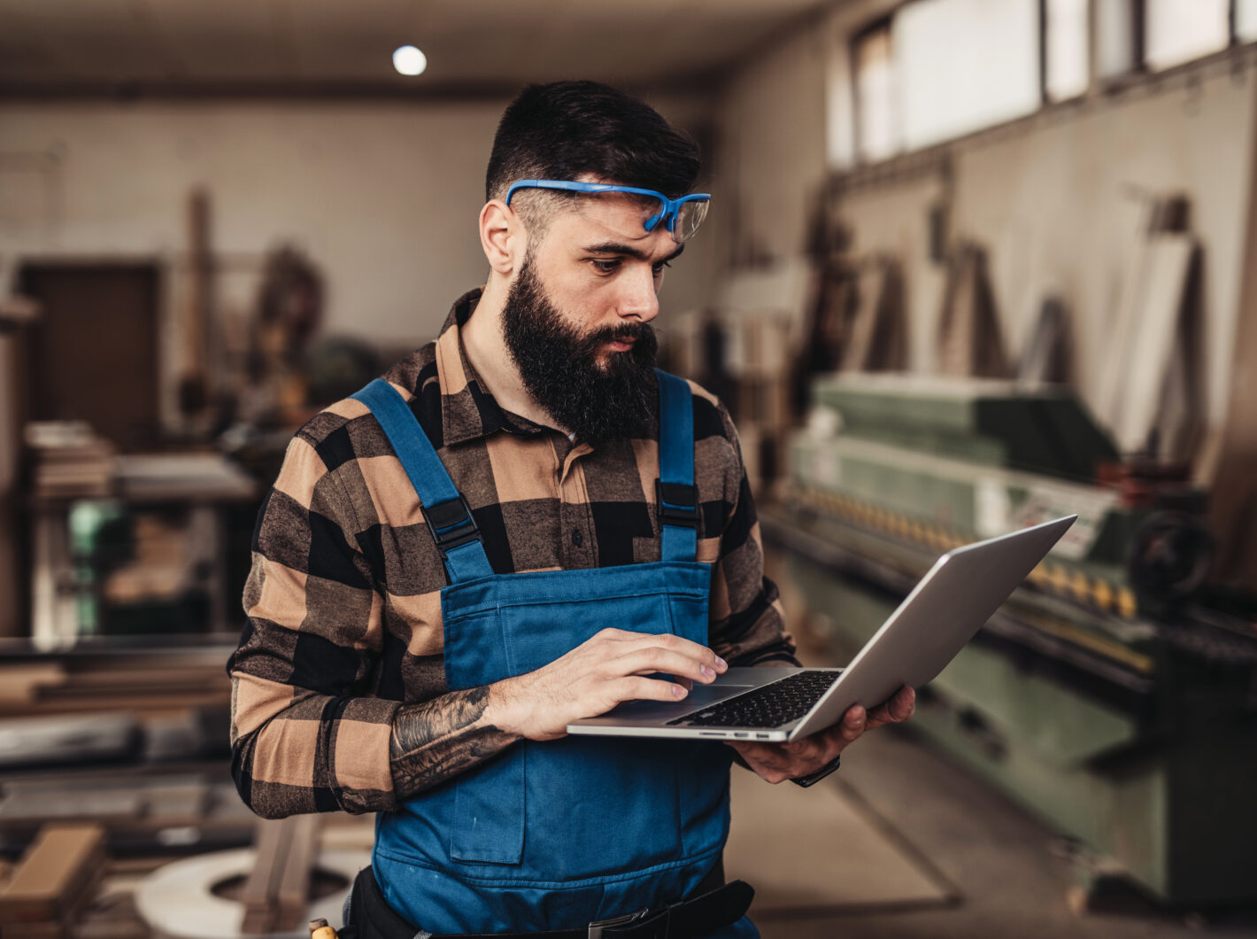 Man in workshop using laptop, wearing blue overalls and goggles.