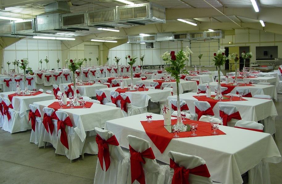 Large banquet hall with tables covered in white linens and red accents, adorned with red and white flower centerpieces.