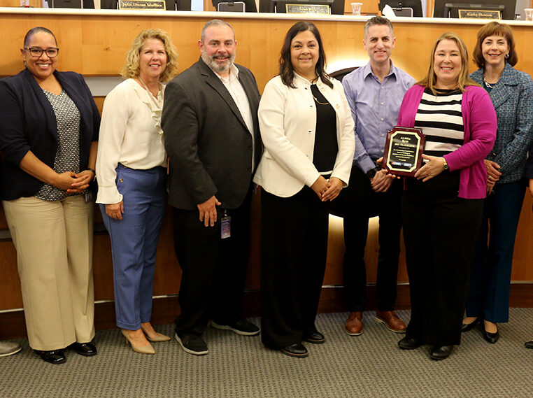 Alisha smiles holding an award plaque, standing with nine colleagues in formal attire.