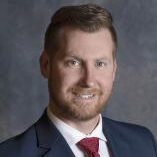 Bill Aiken smiling, wearing a suit and red tie against a gray background