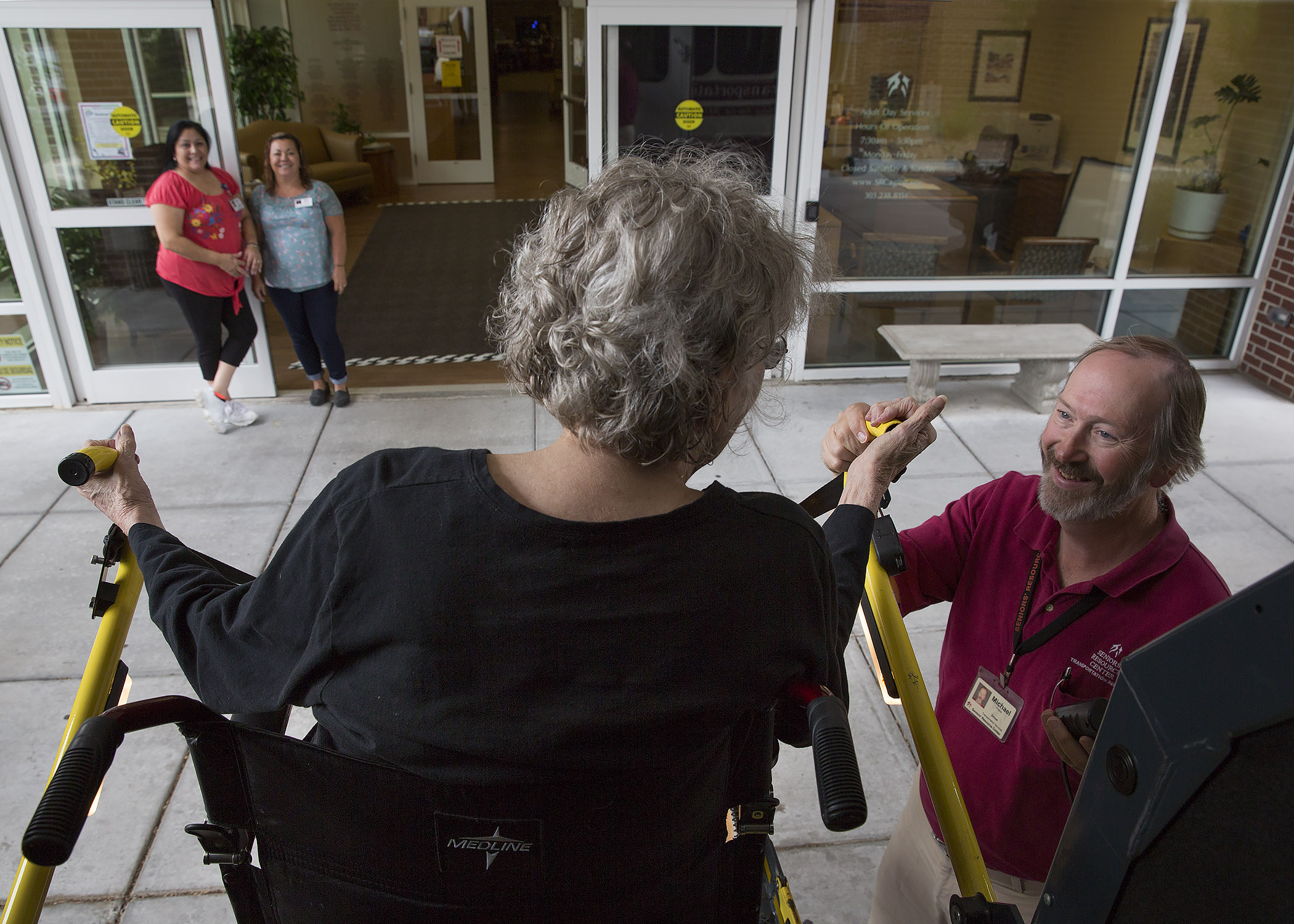 Man assists woman in wheelchair lift; two women watch from entrance.