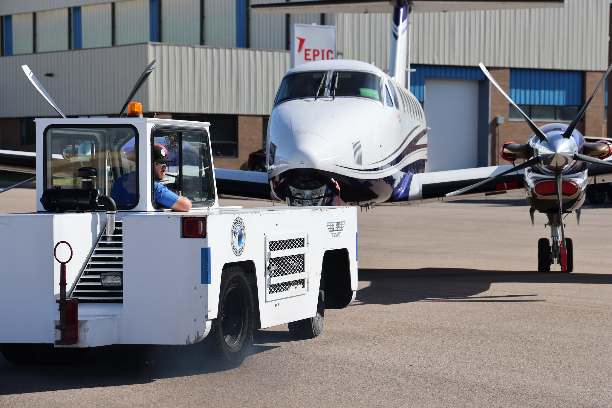 Person towing a plane with a ground vehicle outside a hangar labeled 'Epic'