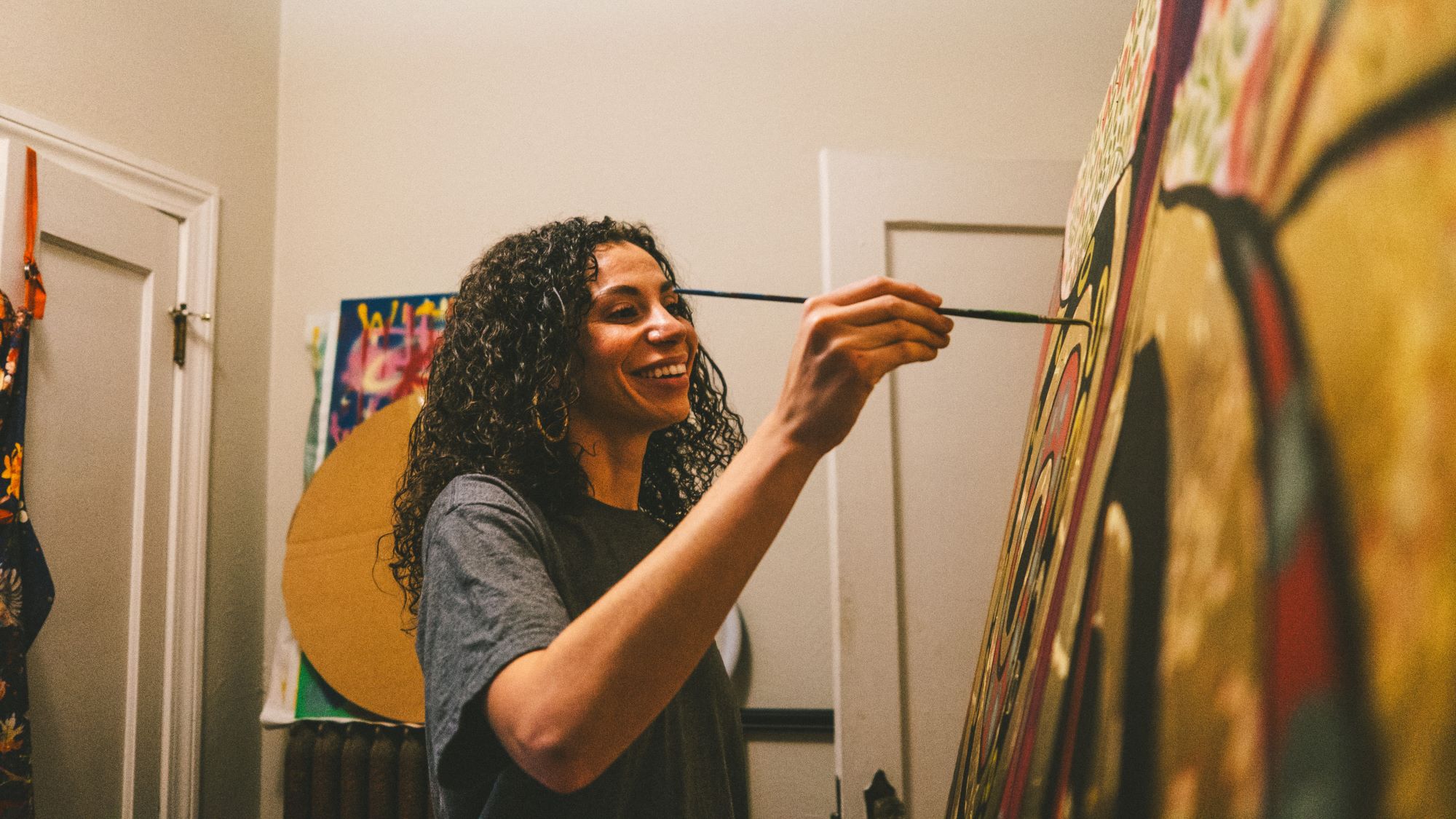 CM8X9596.jpg; person painting a colorful abstract canvas, smiling in a cozy workspace.