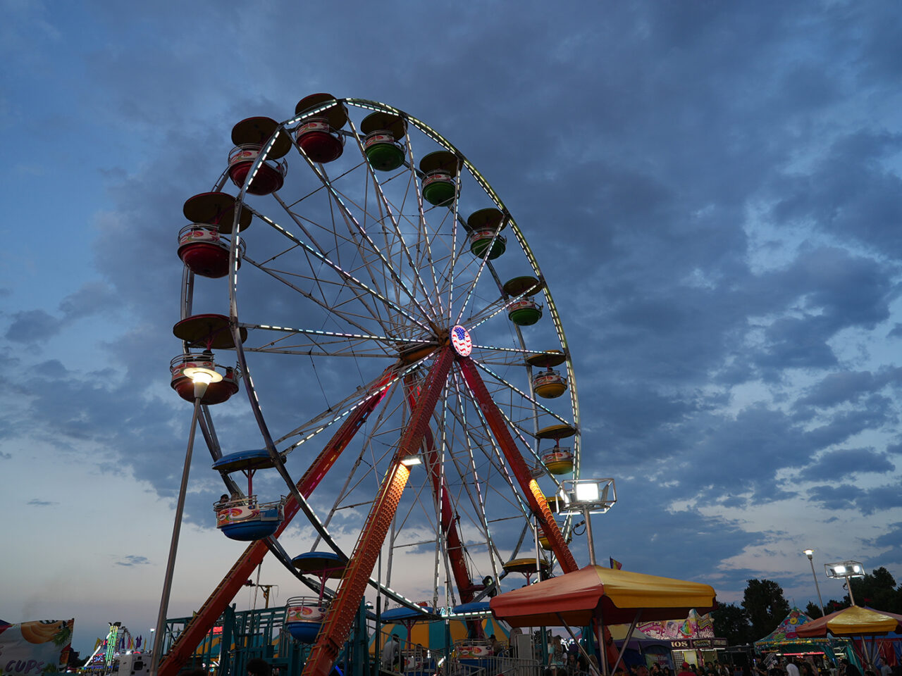 Ferris wheel at dusk with colorful booths and cloudy sky.