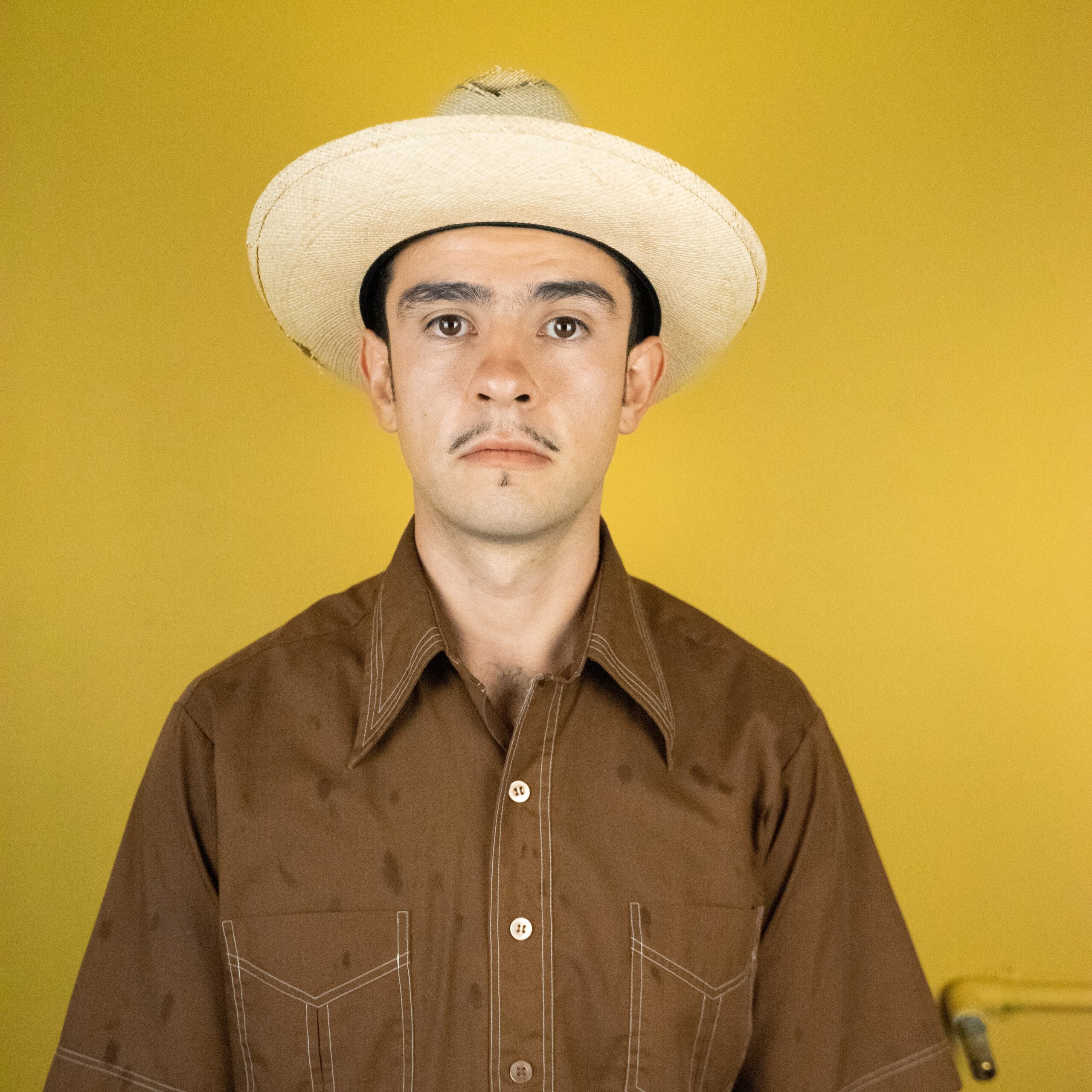 Diego Florez Arroyo wearing a brown shirt and straw hat, neutral expression, yellow background