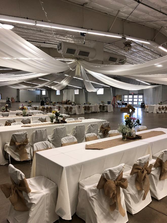 Banquet hall with long tables, white linens, burlap bows, floral centerpieces, and draped ceiling fabric.