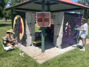 Two artists painting murals on concrete blocks under a pavilion. Sign reads: "Babewalls presents Jahna Rae x Gina McGlash."