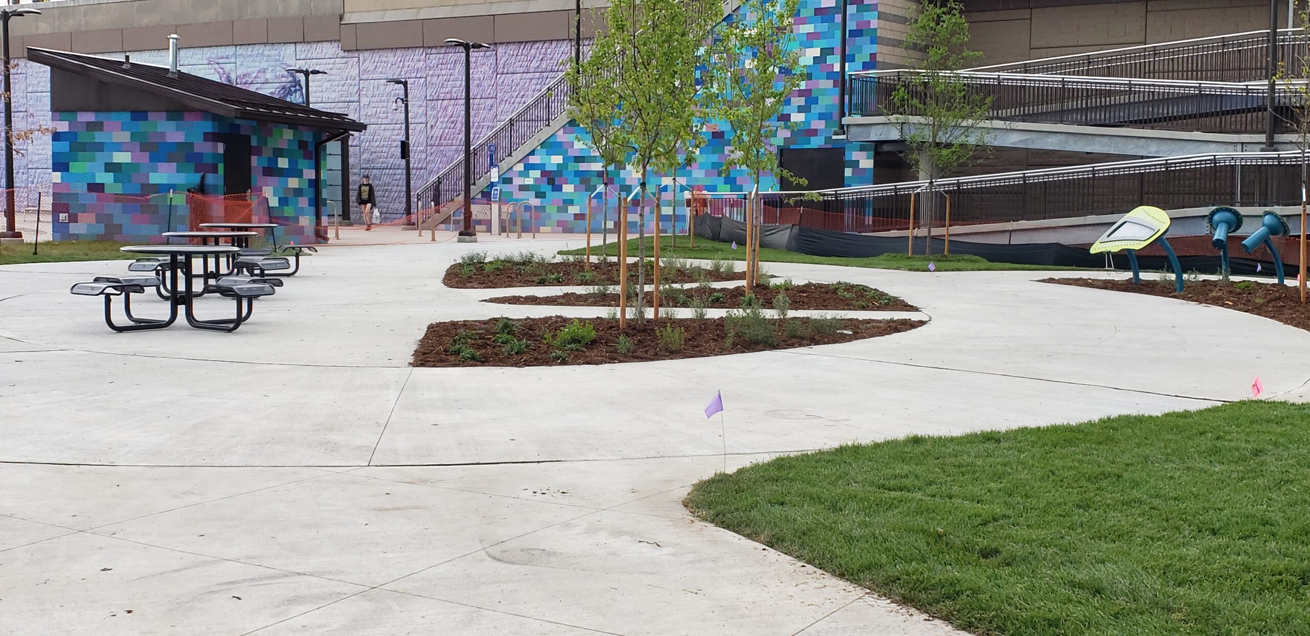 Outdoor plaza with picnic tables, curved walkways, young trees, and colorful tiled building backdrop.