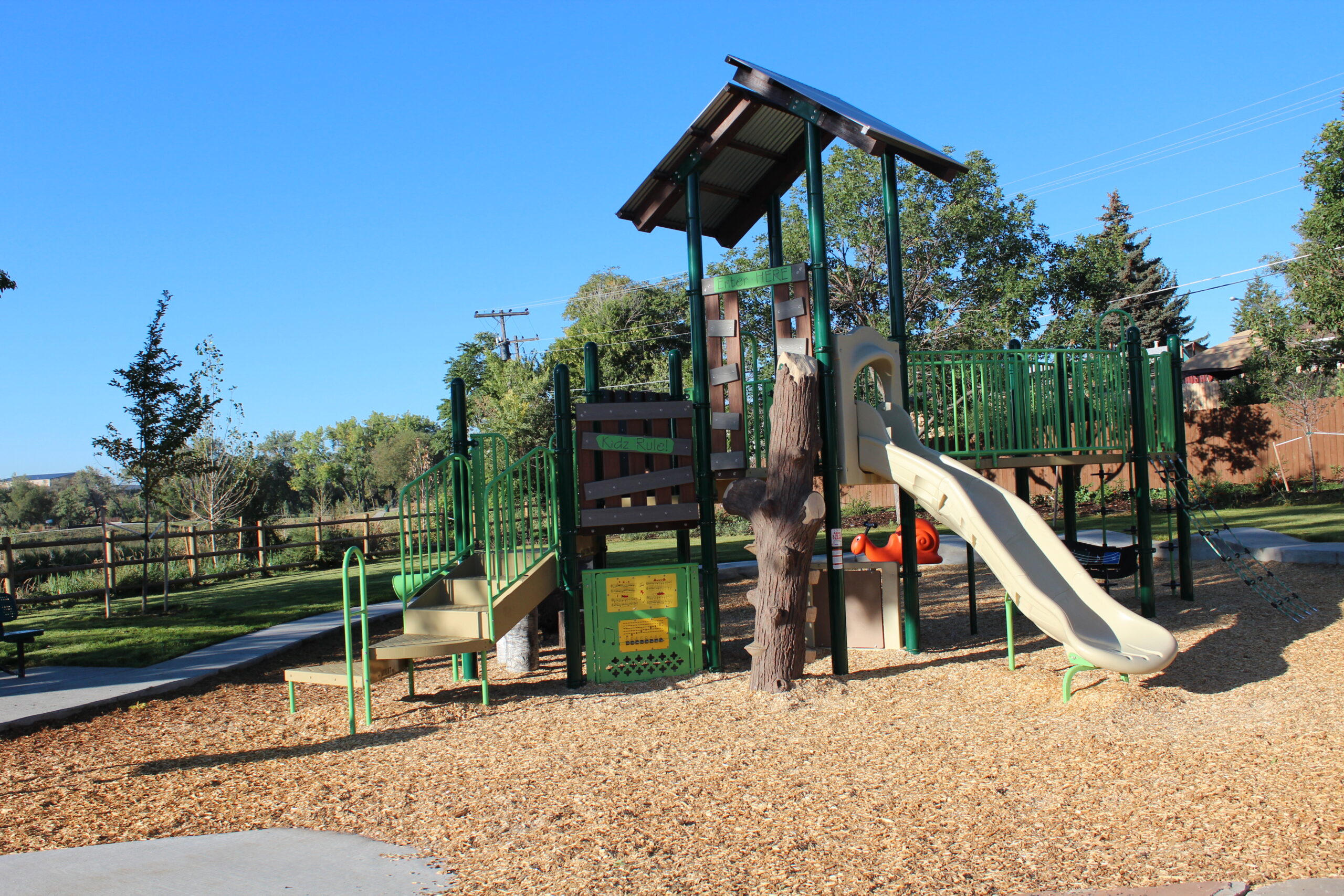 Playground with green climbing structures, beige slide, wood chips, and trees. Signs read "Nidz Rulez" and "Discover Here"