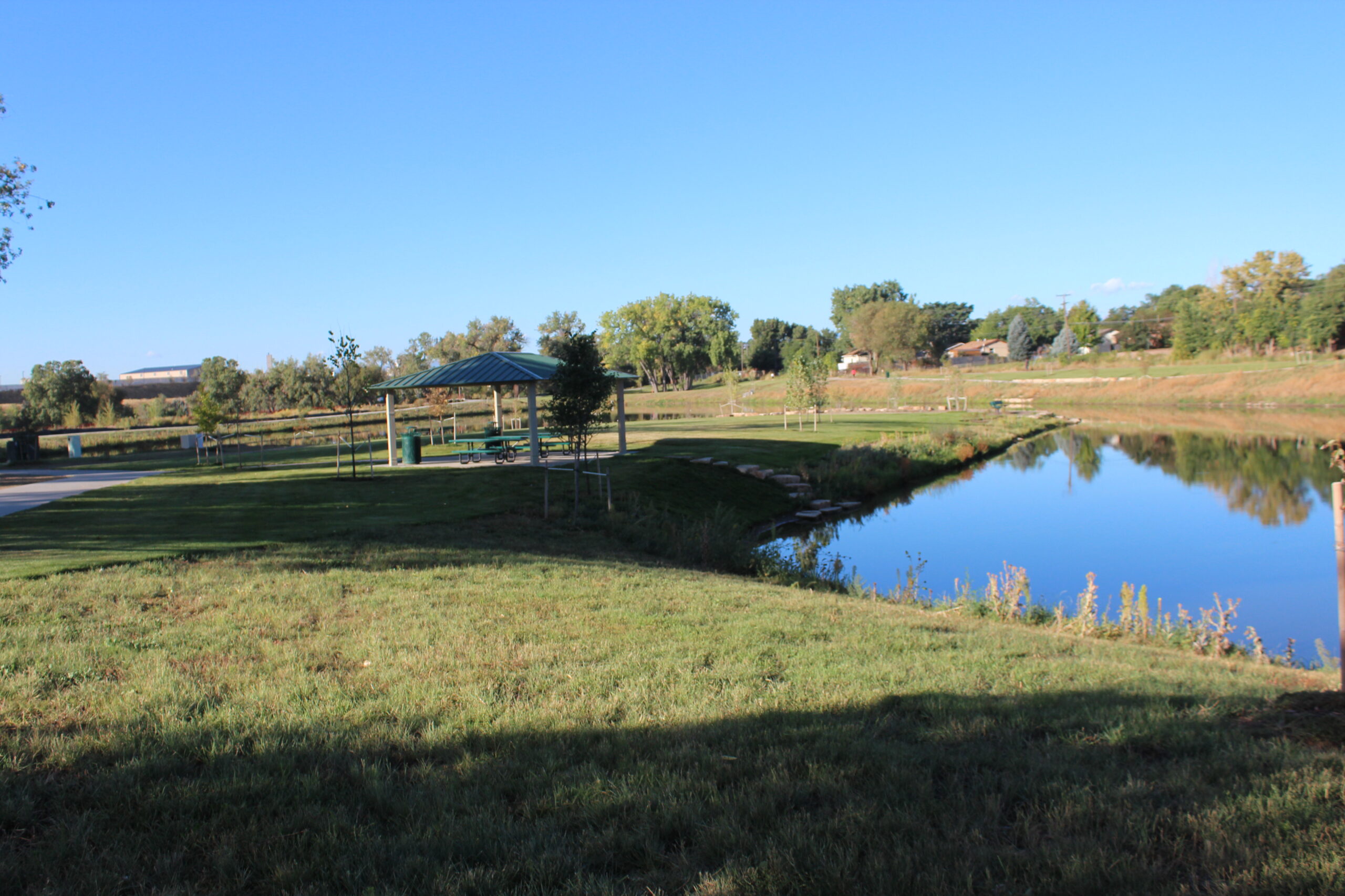 Park scene with gazebo, green lawn, and calm pond reflecting trees.