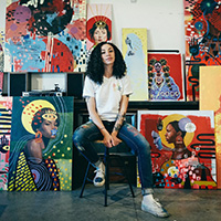 Jahna-Rae sits on a stool, surrounded by vibrant, colorful paintings.