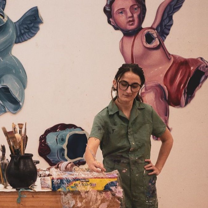 Jenna painting in a studio wearing a green jumpsuit, surrounded by brushes and paint supplies; angel murals on wall