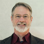 JohnCHicks_web.png smiling with glasses and gray beard, wearing a dark jacket