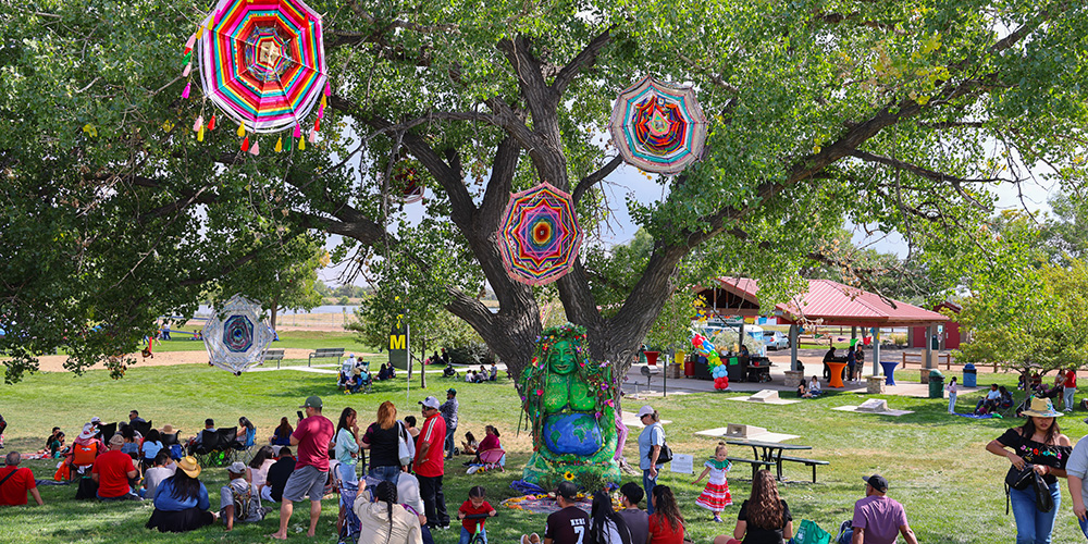 People gathered in a park for a colorful festival, yarn art hanging from trees.