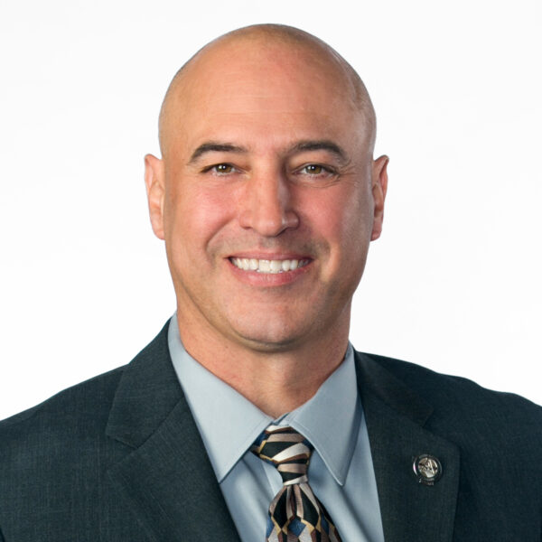 Musso smiling in suit with patterned tie, lapel pin visible