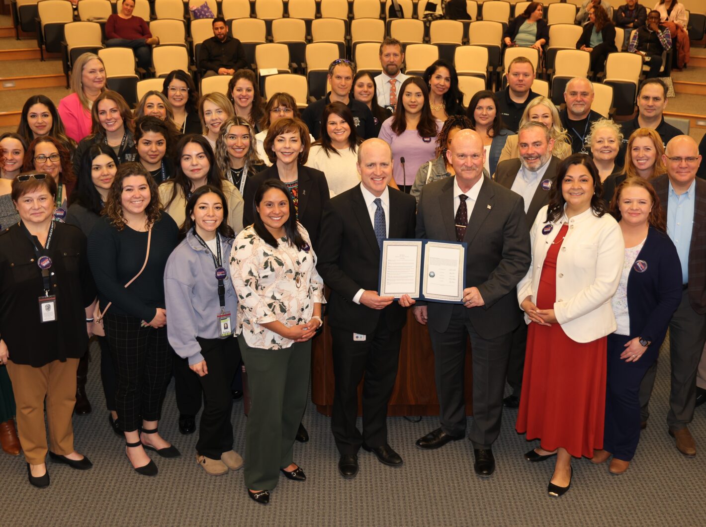 Large group gathered in auditorium; central figures hold an official document recognizing National Crime Victims' Rights Week.