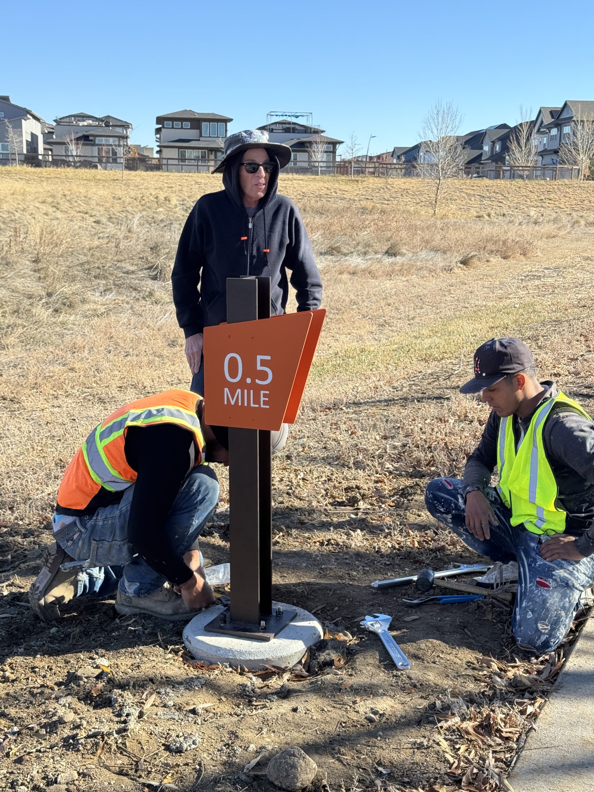 Three workers installing an orange "0.5 Mile" trail marker sign.