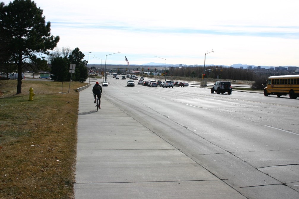 Cyclist on sidewalk beside busy road with cars and school bus in distance