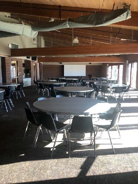 Large conference room with round tables, black chairs, and ceiling beams. Natural light streams through windows.