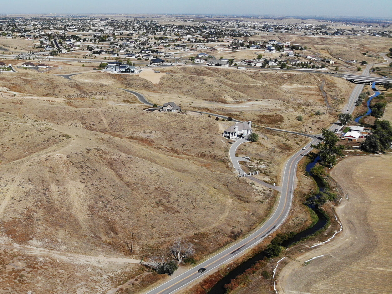 Aerial view of a dry, hilly landscape with scattered houses and winding roads leading to a distant suburban area.