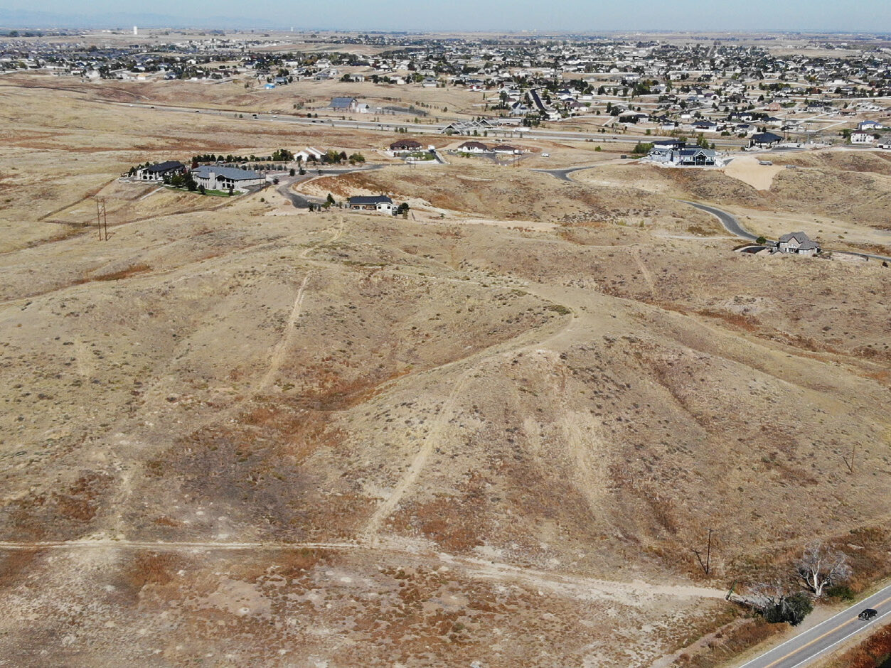 Aerial view of a dry, hilly landscape with scattered houses and winding roads leading to a distant suburban area.
