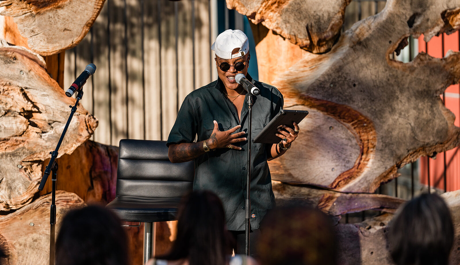 Kerrie Joy sunglasses and cap, speaking into a microphone, holding a tablet.