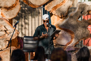 Six.jpg: Person speaking passionately at a microphone, holding a tablet, with an audience and wooden backdrop.