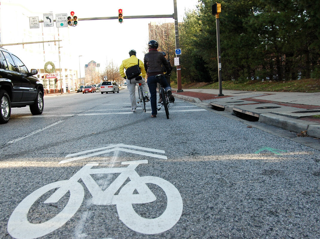 Two cyclists at intersection with visible bike lane marking, next to cars, under a traffic light and signs.