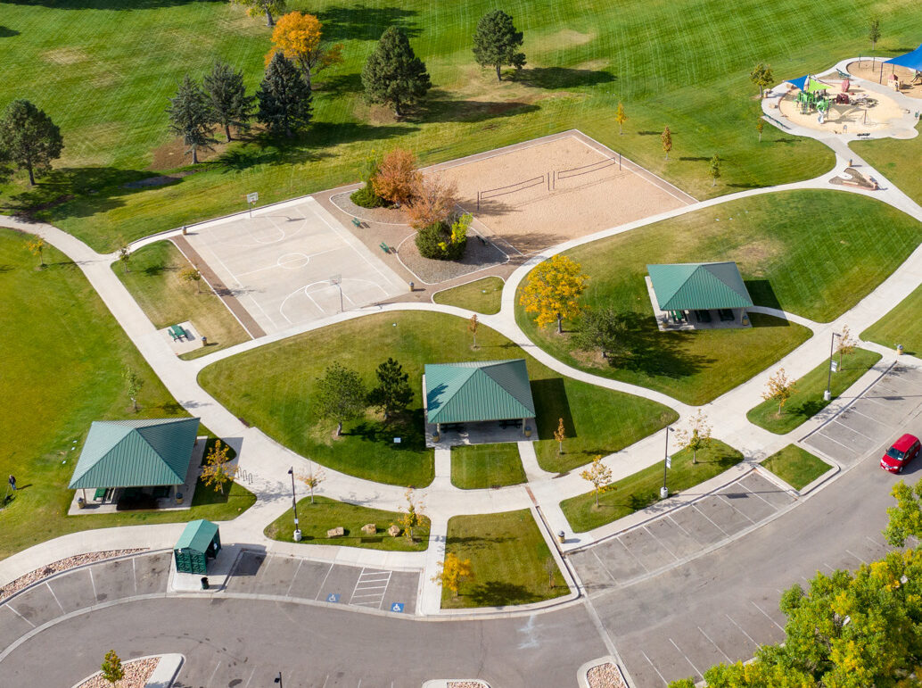 Aerial view of a park with green lawns, pathways, sports courts, and pavilions. Residential area and cityscape in background.