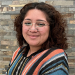 Tania Wilson smiling against a brick wall, wearing glasses and a colorful sweater.