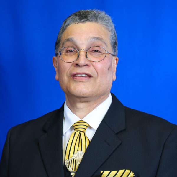 Villagran Alex smiling, wearing glasses, dark suit, yellow striped tie against blue background.