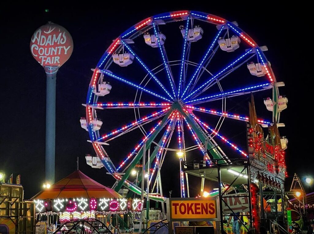 Ferris wheel with colorful lights at night, Adams County Fair sign, illuminated carnival booths, "TOKENS" sign visible.