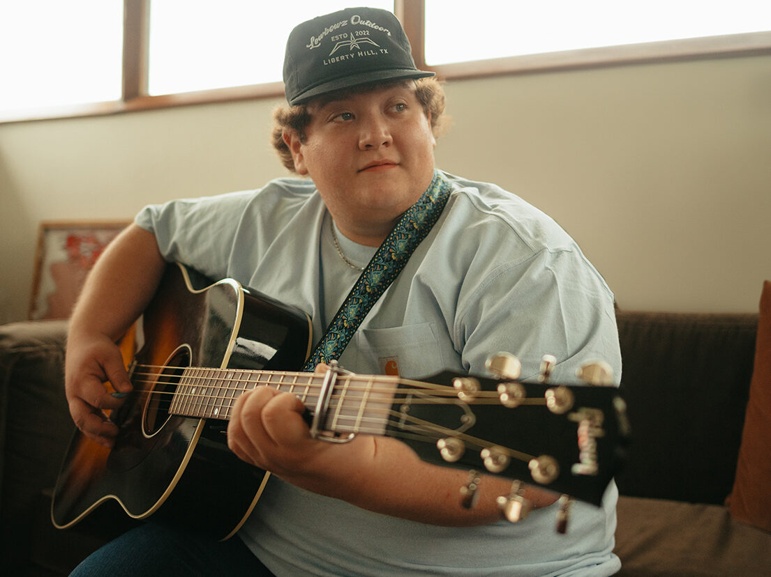 Bayker Blankenship playing guitar, wearing a hat that reads "Legitimus Outdoors ESTD 2022 Liberty Hill, TX"