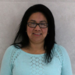 Chavez-1.png: Person wearing glasses, smiling, in blue sweater, standing against a light wall